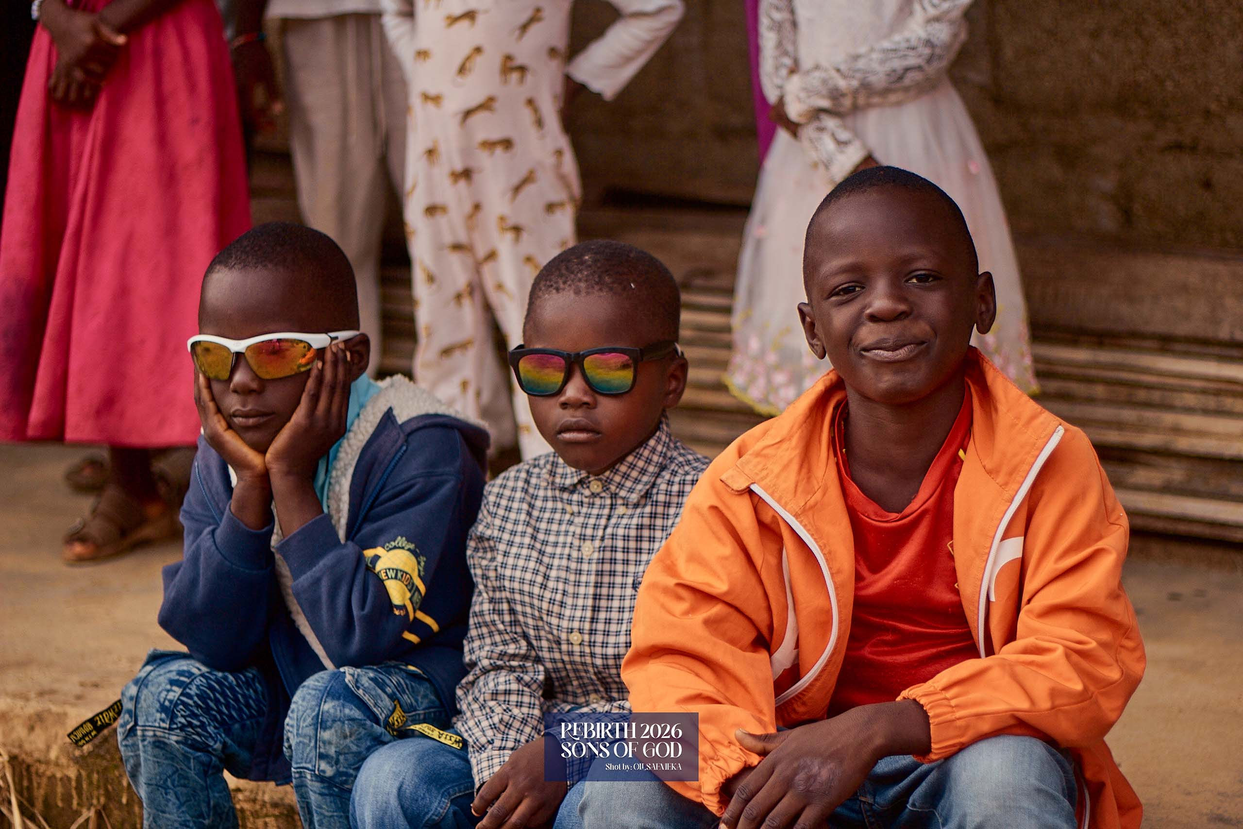 Three children seated together outdoors, two wearing sunglasses.